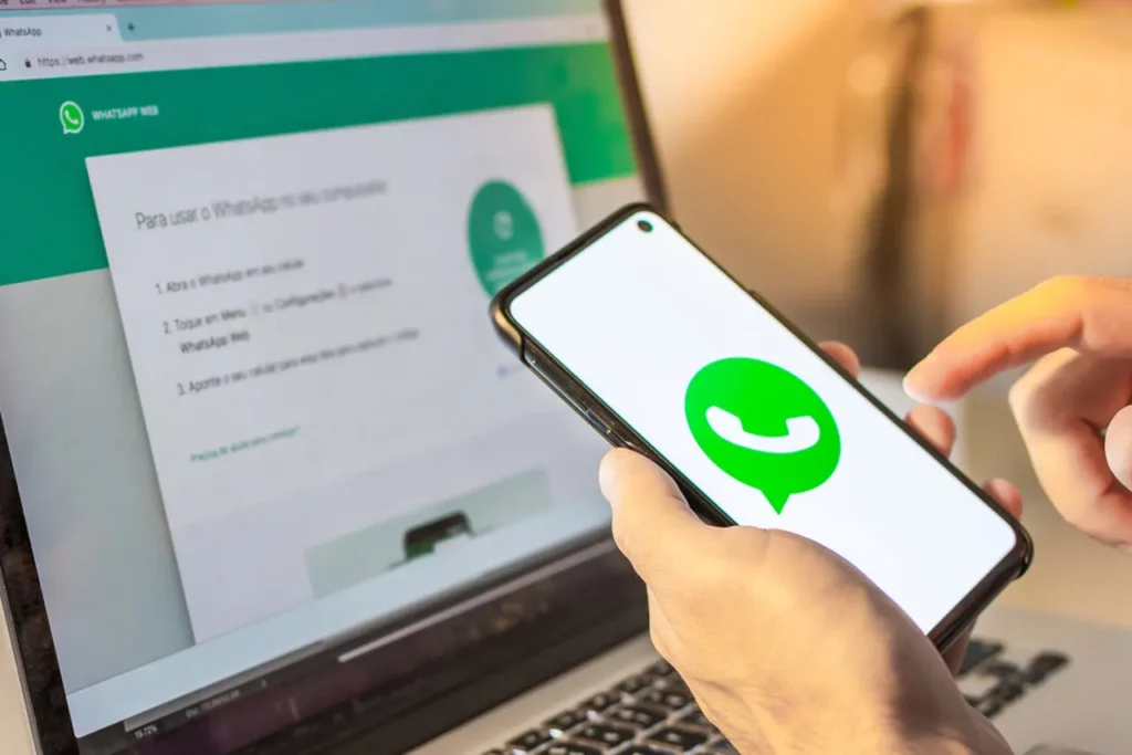 Pessoa com celular na mão com ícone do whatsapp e notebook ao fundo